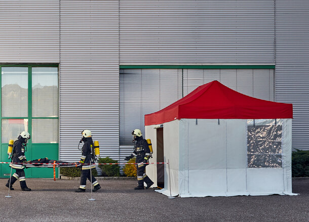 The firemen are carrying the injured person to the rescue tent. The rescue tent has a red roof and grey side walls. 