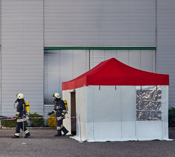 The firemen are carrying the injured person to the rescue tent. The rescue tent has a red roof and grey side walls. 