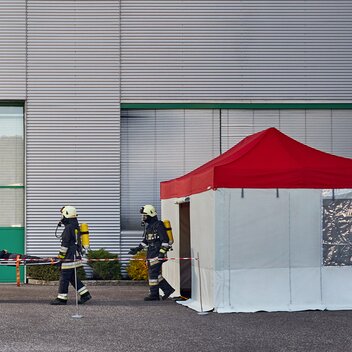 The firemen are carrying the injured person to the rescue tent. The rescue tent has a red roof and grey side walls. 