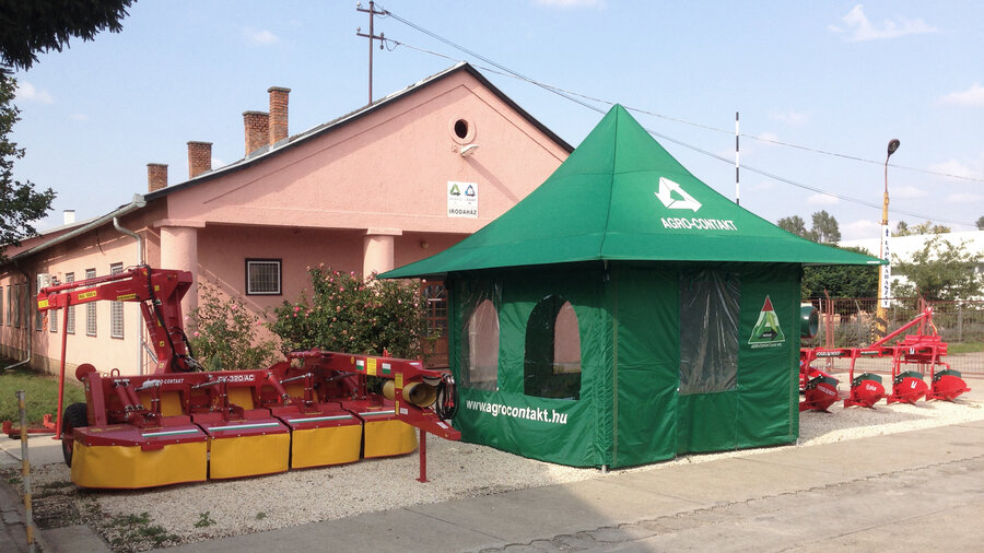 Green agrarian tent equipped with side walls and windows. Next to it there are some agricultural machines.