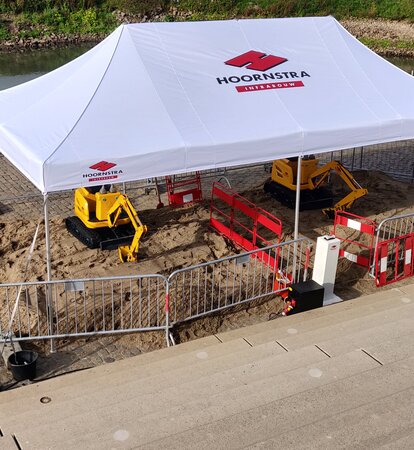 Ein weißer Faltpavillon mit Logo-Aufdruck "HOORNSTRA" wird zur Überdachung der Baustelle genutzt. Darunter arbeiten zwei gelbe Bagger an der Straßenreparatur.