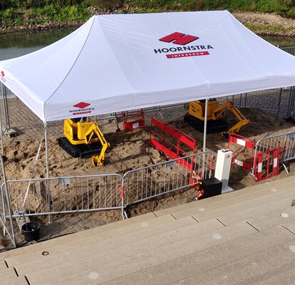 A white gazebo with the logo imprint "HOORNSTRA" is roofing the construction site. Underneath, two yellow excavators are working on the road maintenance. 