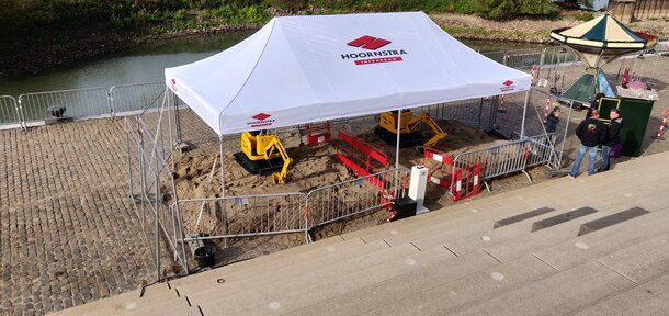 A white gazebo with the logo imprint "HOORNSTRA" is roofing the construction site. Underneath, two yellow excavators are working on the road maintenance. 