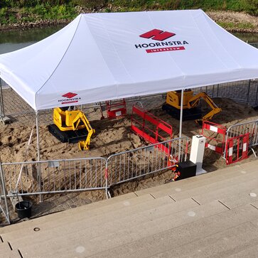 A white gazebo with the logo imprint "HOORNSTRA" is roofing the construction site. Underneath, two yellow excavators are working on the road maintenance. 