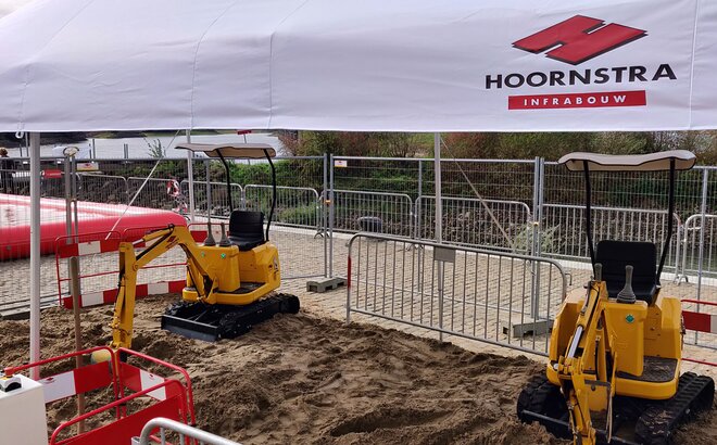 A white gazebo with the logo of "Hoornstra" is covering a construction site with yellow excavators standing on a sandy ground.