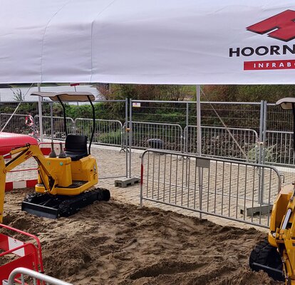 Un gazebo pieghevole con la scritta "Hoornstra" copre un cantiere con delle ruspe gialle su terreno sabbioso.