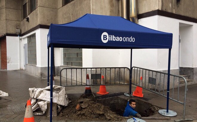 Ein blauer Faltpavillon mit dem Firmenlogo "Bilbao ondo" überdacht einen Bauarbeiter, der ein Loch gräbt.