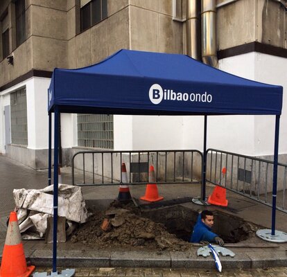 Un gazebo pieghevole blue con il logo aziendale "Bilbao ondo" copre un operaio che sta scavando un buco. 