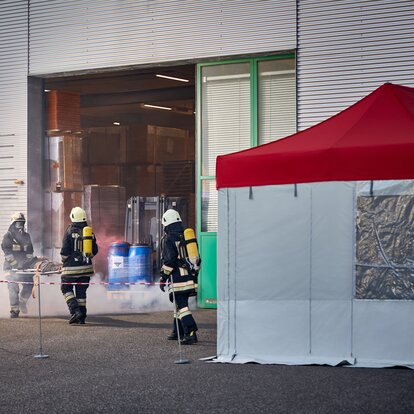 The firemen are carrying a victim of a chemical accident out of the hall. They are bringing the injured person to the gazebo. 