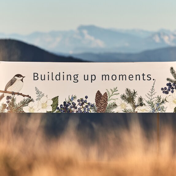 The white folding gazebo with flat roof is printed with forest motifs such as birds, pine cones and leaves. Above it is written "building up moments".