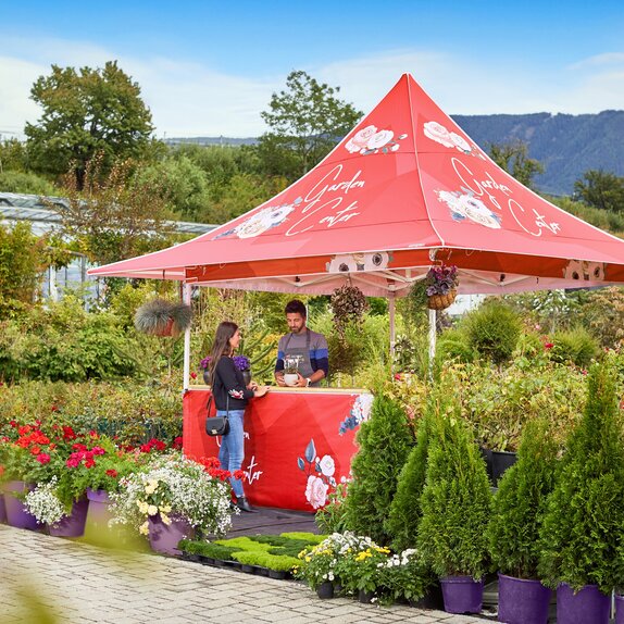 Ein mit Blumen bedruckter Faltpavillon mit Vordach steht inmitten verschiedenster Pflanzen einer Gärtnerei. Der Verkäufer im Faltpavillon spricht mit der Verkäuferin.