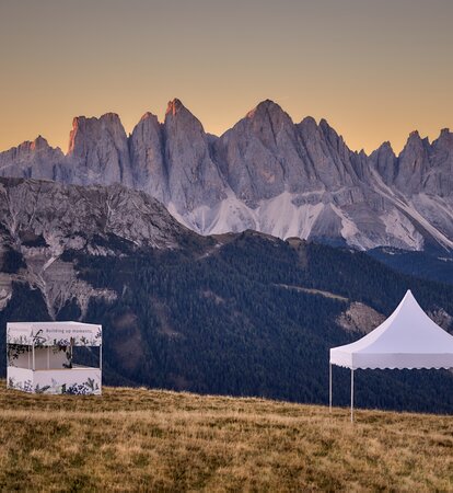 Ein bedrucktes weißes Faltzelt mit Flachdach und ein weißes Pagodenzelt stehen auf dem Plose-Berg. Die beiden Faltpavillons stehen im Sonnenuntergang vor einer Bergkette.