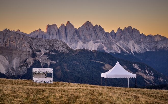 Ein bedrucktes weißes Faltzelt mit Flachdach und ein weißes Pagodenzelt stehen auf dem Plose-Berg. Die beiden Faltpavillons stehen im Sonnenuntergang vor einer Bergkette.