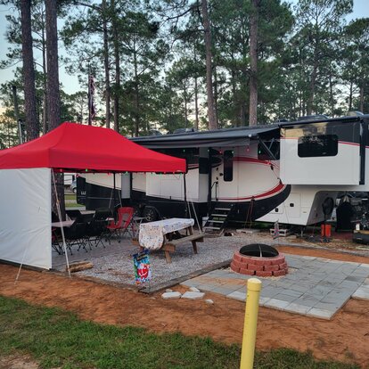 Camping-car avec barnum pliante intégrée dans la forêt. Sous la barnum se trouve une table de camping avec des chaises, entourée de grands arbres.