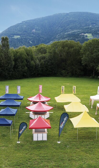 Alle Faltzelte in allen Größen und Ausführungen stehen nach Farbe getrennt auf einer großen Wiese. Die Faltpavillons sind entweder blau, rot, gelb oder in ecru.