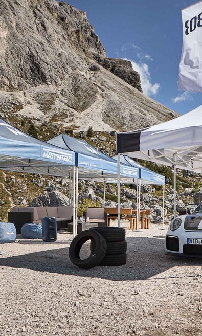 Next to a road there are three blue and one white folding gazebo with black print. Under the white tent there is a racing car and mountains can be seen in the background.