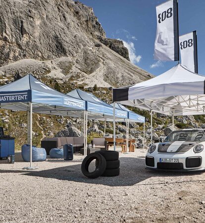 Accanto a una strada ci sono tre gazebo pieghevoli blu e uno bianco con stampa nera. Sotto il gazebo bianco c'è una macchina da corsa e sullo sfondo si vedono delle montagne.