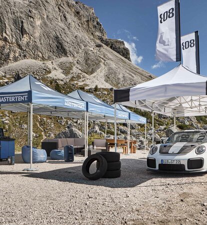 Accanto a una strada ci sono tre gazebo pieghevoli blu e uno bianco con stampa nera. Sotto il gazebo bianco c'è una macchina da corsa e sullo sfondo si vedono delle montagne.