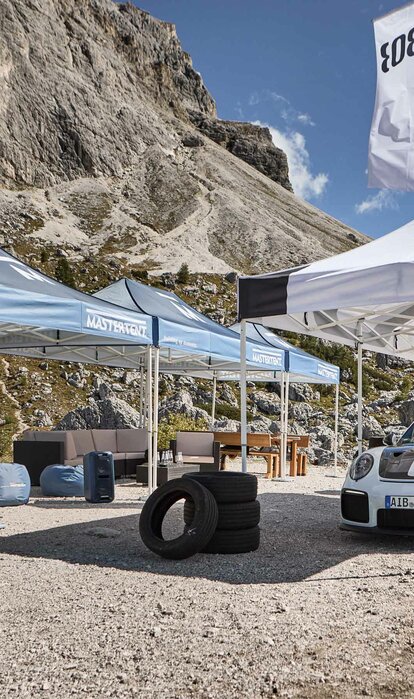 Next to a road there are three blue and one white folding gazebo with black print. Under the white tent there is a racing car and mountains can be seen in the background.