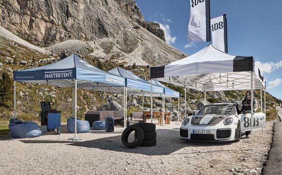 Accanto a una strada ci sono tre gazebo pieghevoli blu e uno bianco con stampa nera. Sotto il gazebo bianco c'è una macchina da corsa e sullo sfondo si vedono delle montagne.