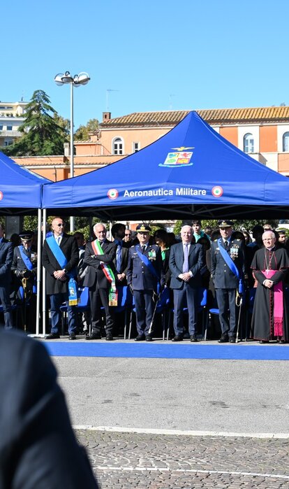 Veranstaltung der italienischen Luftwaffe im Freien. Unter drei blauen Faltpavillons stehen Vertreter aus Militär. Im Vordergrund sind zwei uniformierte Personen von hinten zu sehen.