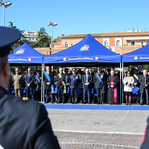 Veranstaltung der italienischen Luftwaffe im Freien. Unter drei blauen Faltpavillons stehen Vertreter aus Militär. Im Vordergrund sind zwei uniformierte Personen von hinten zu sehen.