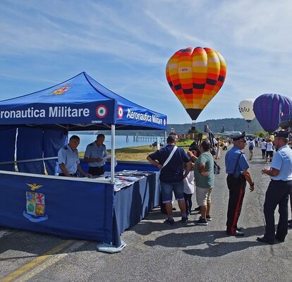 Informationsstand der italienischen Luftwaffe am Ufer, umgeben von Besuchern und Polizisten. Im Hintergrund steigen bunte Heißluftballons in den Himmel.
