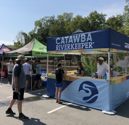Ein Promotionszelt der Catawba Riverkeeper Foundation steht am Straßenrand. Das Square-Zelt ist vollbedruckt und hat eine Holztheke. Ein Mitarbeiter spricht mit einer Frau vor der Theke.