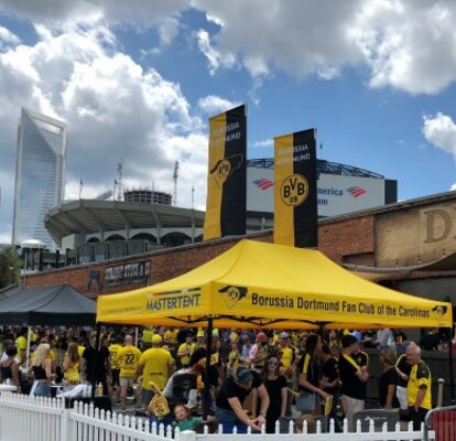 Ein gelber Pavillon des BVB Borussia Dortmund Fan Clubs steht hinter einem weißen Zaun. Er hat zwei große banner auf dem Dach. 