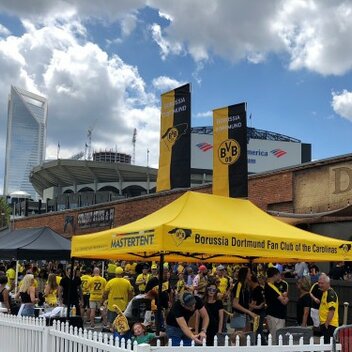 Ein gelber Pavillon des BVB Borussia Dortmund Fan Clubs steht hinter einem weißen Zaun. Er hat zwei große banner auf dem Dach. 