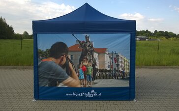 A dark blue 3x3 m promotion tent stands outside on a paved path. The complete side wall is printed with a photo. The photo shows a family taking a photo in front of a statue. 