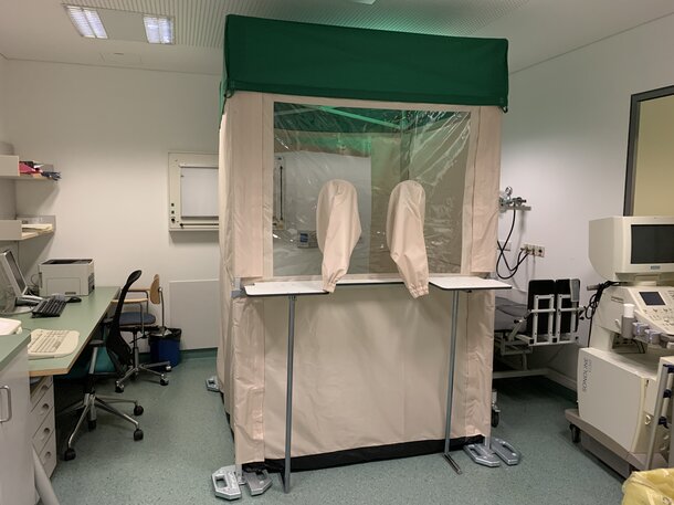 A 1,5x1,5 m Covid-19 test cabin is located in the hospital in Schlanders. The test cabin is equipped with gaiters and a protective wall. The test cabin also has a green flat roof. Next to the test cabin on the left side is a desk with a chair and on the right side a device. 