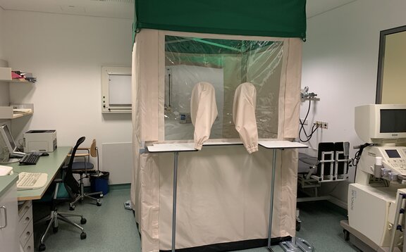 A 1,5x1,5 m Covid-19 test cabin is located in the hospital in Schlanders. The test cabin is equipped with gaiters and a protective wall. The test cabin also has a green flat roof. Next to the test cabin on the left side is a desk with a chair and on the right side a device. 