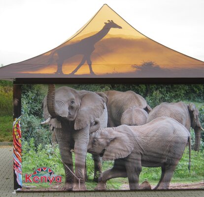 Una tenda stampata 4x4 m con 4 tettoie dalla parte anteriore. Sul tetto si vede una giraffa al tramonto e sulla parete laterale ci sono diversi elefanti. Sulla parete laterale in basso a sinistra si vede anche il logo "Kenya" nel colore rosso. 