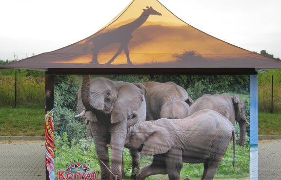 A 4x4 m printed tent with 4 canopies from the front. On the roof is a giraffe in the sunset and on the side wall are several elephants. On the side wall on the lower left you can see the logo "Kenya" in red. 