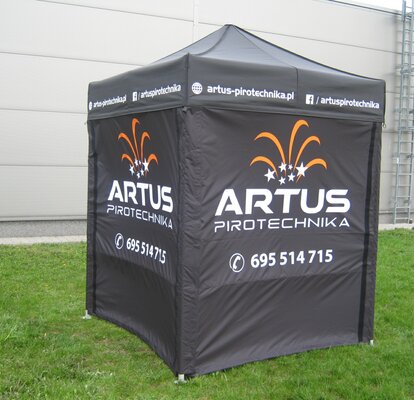 A black 2x2 m promotion tent. The side walls are printed with the logo "Artus Pirotechnika" in white and partly orange, as well as with the telephone number. The tent stands on the meadow. 