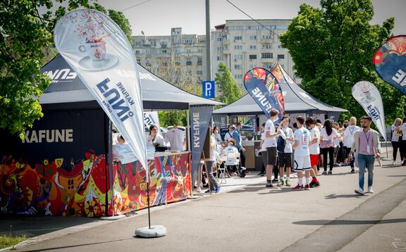 3x3 m-es összecsukható pavilon egyedi nyomtatással és Kafune logóval, teljes oldalfalakkal, félmagas oldalfalakkal, pultokkal és egyedi zászlókkal. A pavilonokat 28 kg-os súlyokkal rögzítik a talajhoz.