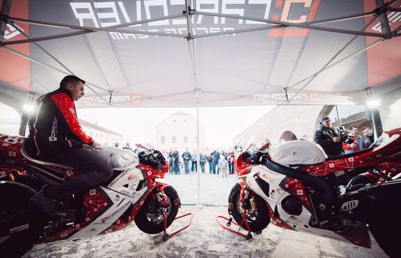 In the tent there are two motorcycles, each in the colours white, black and red. A driver sits on the left side of a motorcycle.