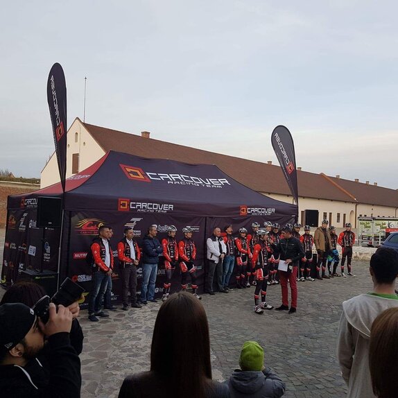 An 8x4 m tent with closed printed side walls in the colour dark blue. The tent is printed with the logo "Carcover Racing Team". On the roof left and right are printed flags. The whole team is standing in front of the tent. 