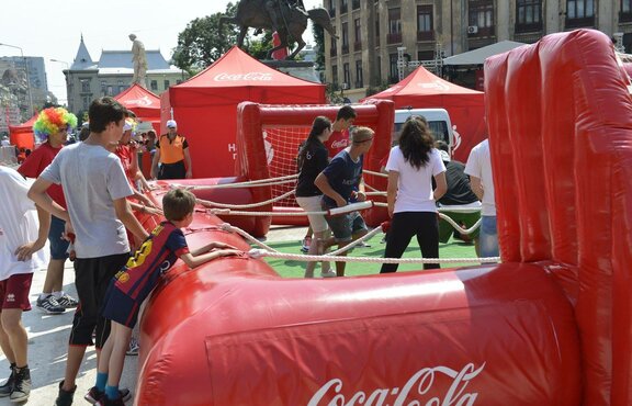 A few children play in a fenced bouncy castle-like football field, which is red and printed with the logo "Coca Cola". In the background you can see a red Coca Cola tent. 