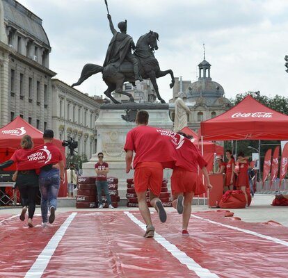 2 Paare tragen jeweils zusammen 1 T-Shirt und machen ein Wettrennen. Dahinter sieht man links und rechts ein 3x3 m rotes Zelt mit dem Logo "Coca Cola" auf dem Dach bedruckt. Sowie in der Mitte steht ein große Statue - ein Mann auf einem Pferd. 