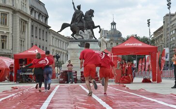 2 couples wear 1 t-shirt together and have a race. Behind it you see left and right a 3x3 m red tent with the logo "Coca Cola" printed on the roof. In the middle there is a big statue - a man on a horse. 