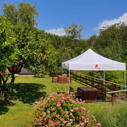 Una tenda di 3x3 m con tetto stampato bianco con il logo Autism Aid. La tenda si trova all'aperto su un prato tra gli alberi. Un tavolo da giardino con sedie è sistemato sotto la tenda.