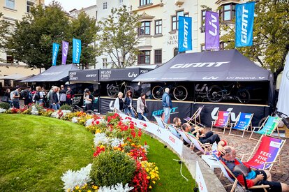 Auf dem Bild sind zwei Faltpavillons mit Flachdach und zwei Faltpavillons mit Spitzdach und Vordach zu sehen. Sie haben zudem Flaggen auf dem Dach. Alle Faltpavillons haben eine Theke und sind personalisiert.