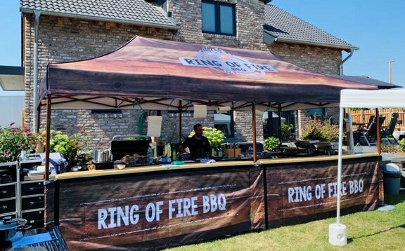 Der Faltpavillon von Ring of Fire BBQ steht im Grünen vor einem Haus. Der Faltpavillon ist vollflächig bedruckt.