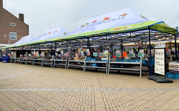 I gazebo per mercati di Vitamientje.nl sono uno accanto all'altro. Hanno una tettoia e sono completamente stampati. Sotto di loro ci sono le bancarelle di frutta.