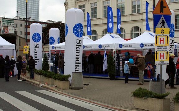 I tre gazebo promozionali bianchi di Skandia si trovano sulla piazza. Tutt'intorno a loro, la gente sta raccogliendo informazioni. Ci sono bandiere blu sui tetti dei gazebo promozionali.