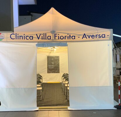 The rescue tent of the hospital "Clinica Villa Fiorita" stands in front of the hospital in the evening. Lights are burning inside.