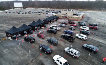 There are 8 black tents in a parking lot in the USA. Cars drive through the testing station next to a building.
