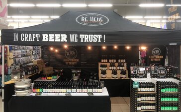 A black folding gazebo with the writing In craft beer we trust is standing in a supermarket. Different beer types of the Be Here brewery are displayed.